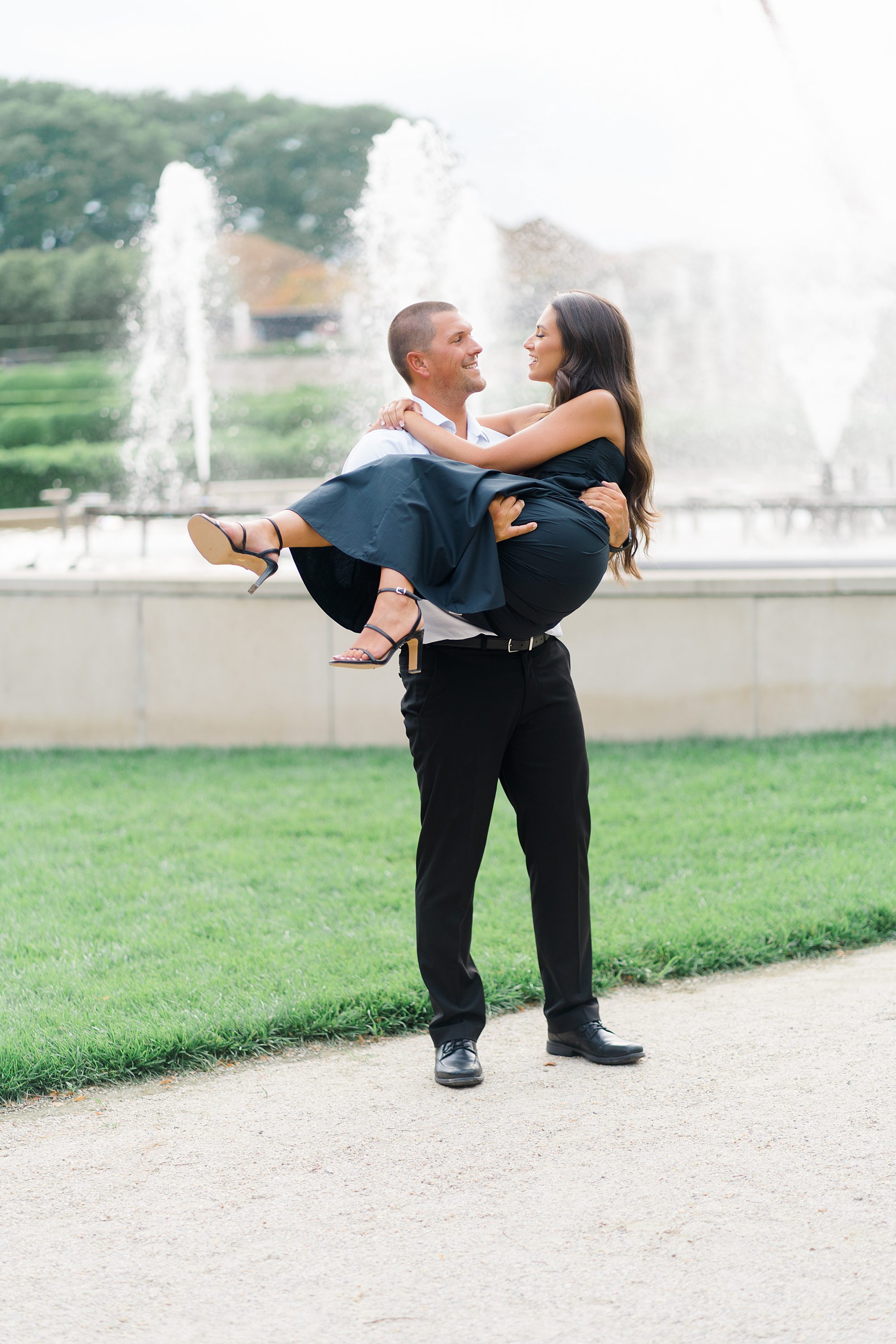 man lifts his fiancé up in front of fountain
