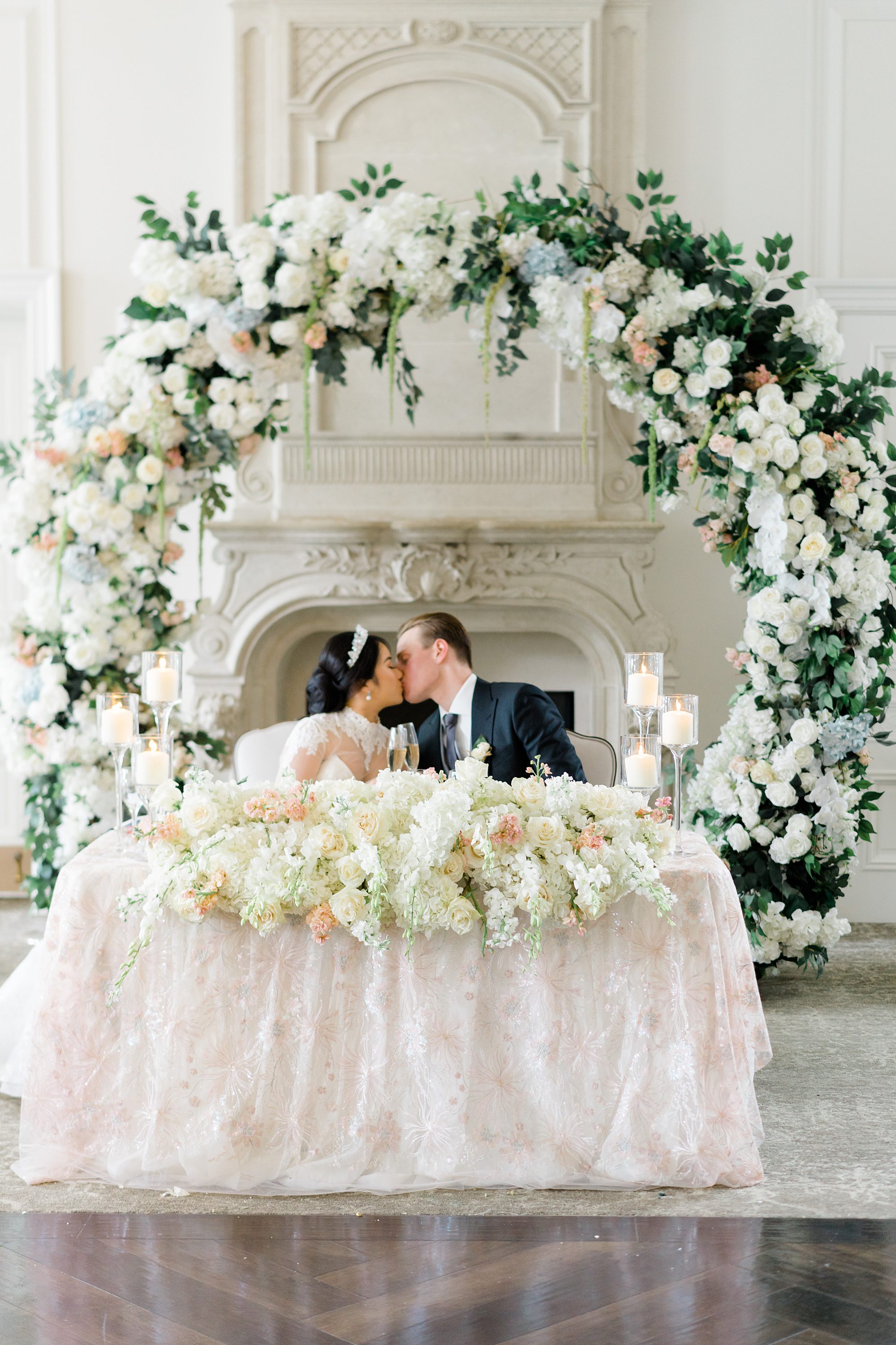newlyweds kiss sitting at their sweetheart table under a flower arch