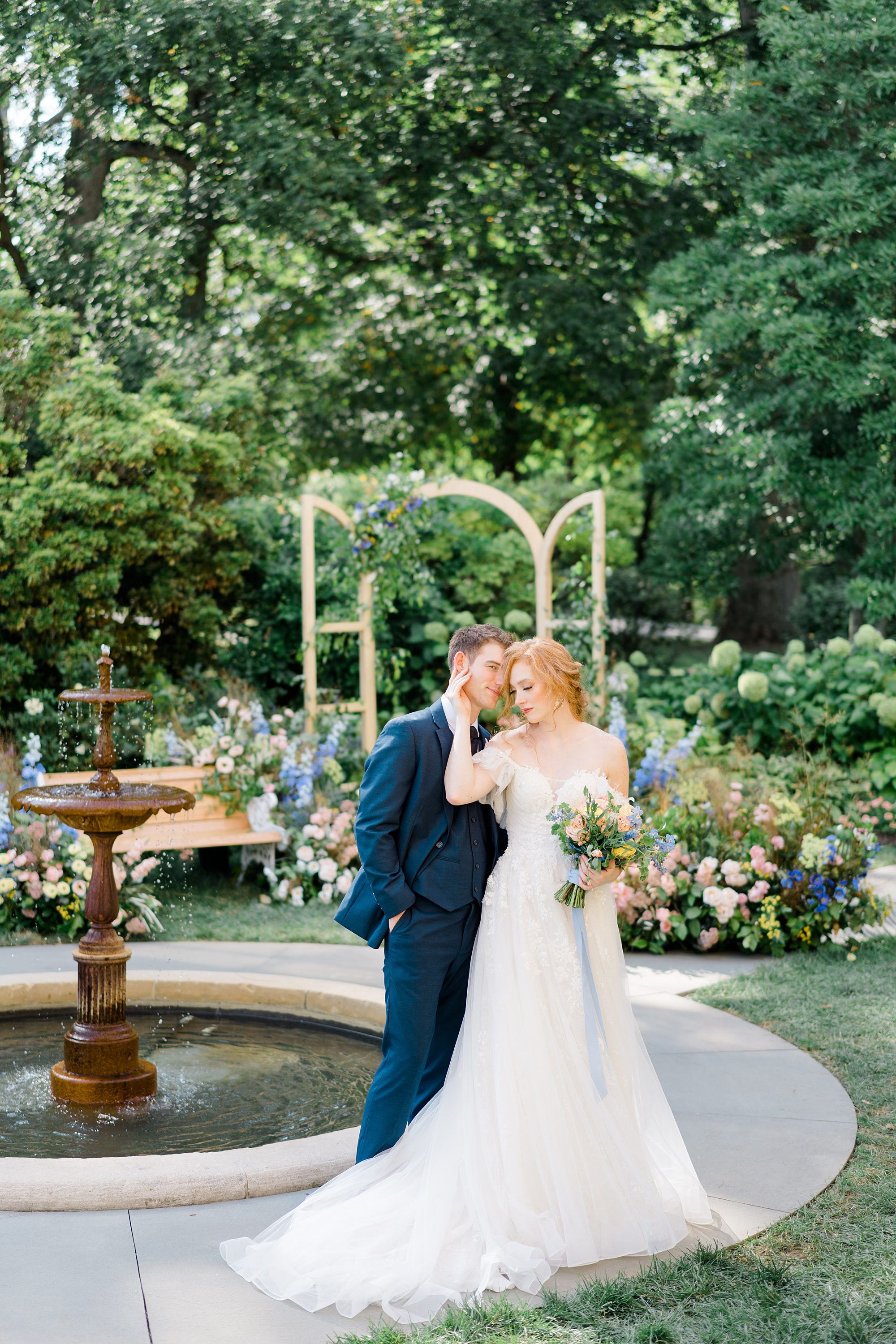 bride holds her hand to grooms face as they stand in front of the water fountain