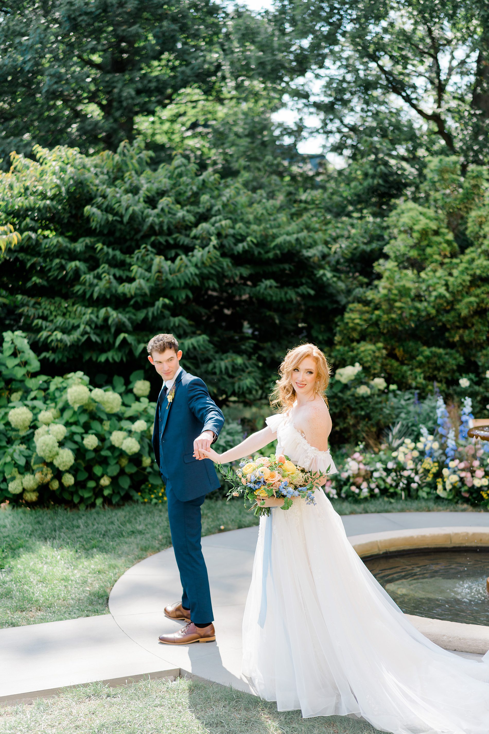 groom leads bride around water fountain in garden of Cairnwood Estate