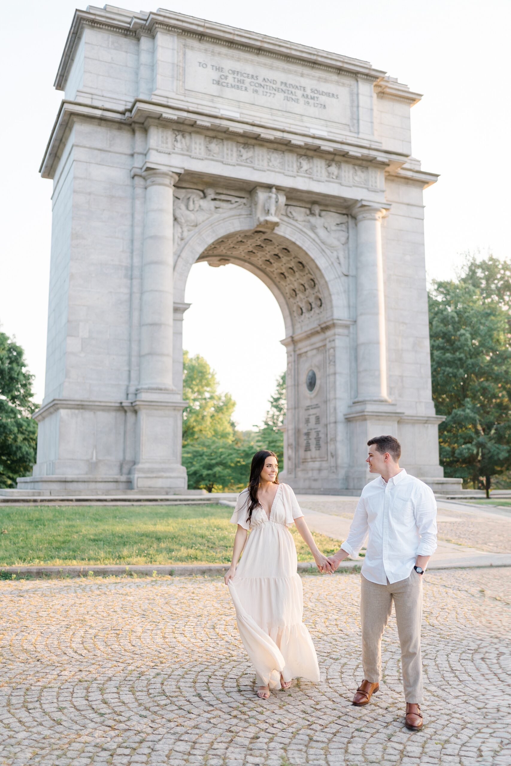 engaged couple hold hands as they walk in front of the National Memorial Arch at Valley Forge, PA