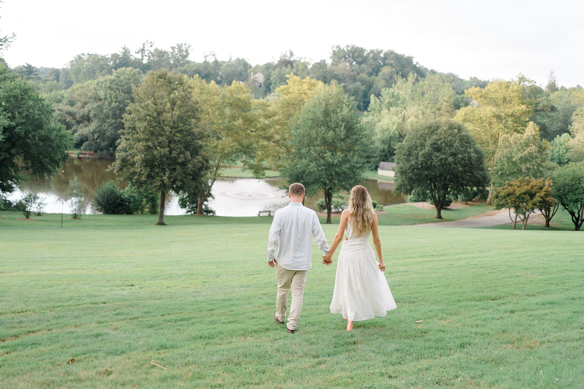 Engaged couple walk towards the trees and pond at The Willows