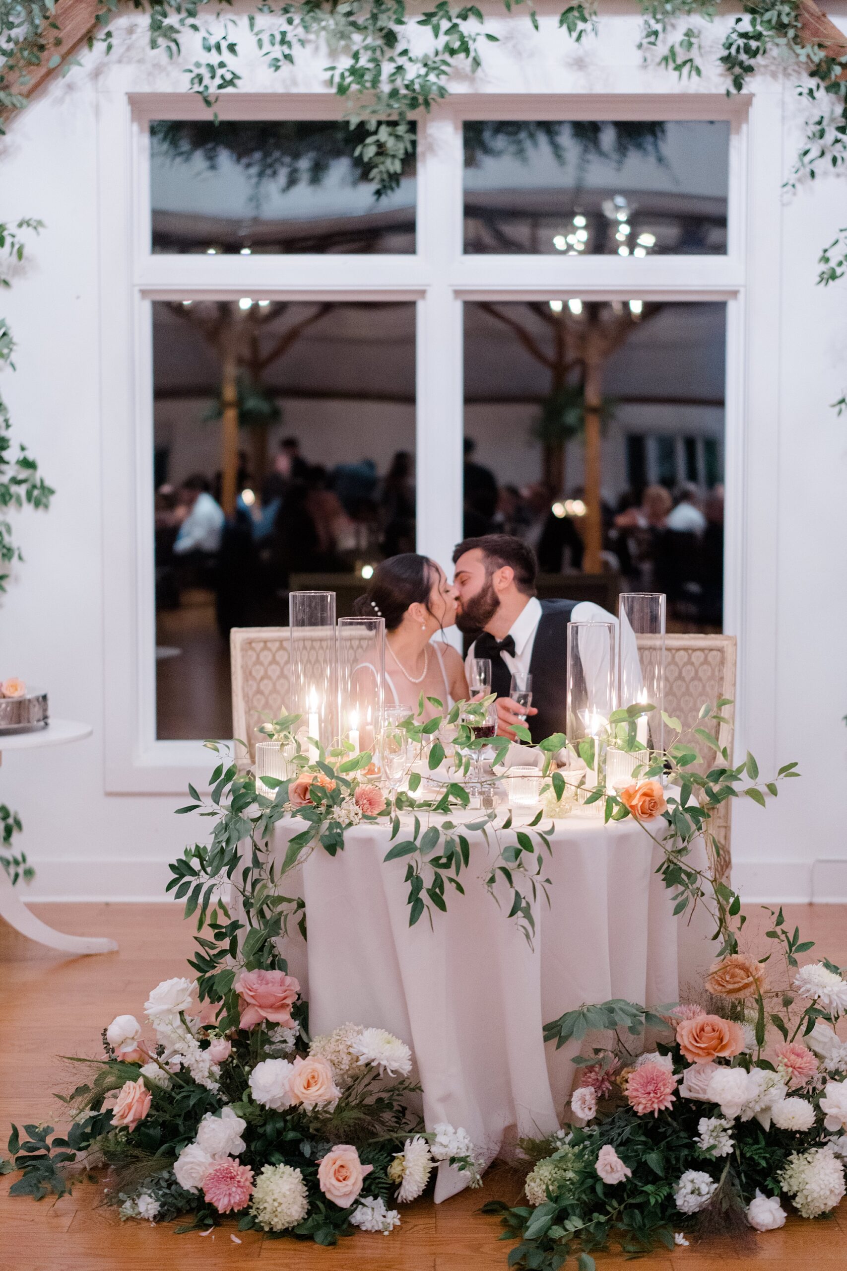 newlyweds kiss as they sit together at Romantic Floral-Centered Wedding reception