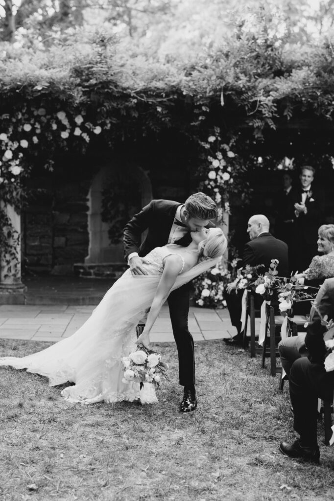 Black and white image of bride and groom kissing as they walk down the aisle at Curtis Arboretum wedding